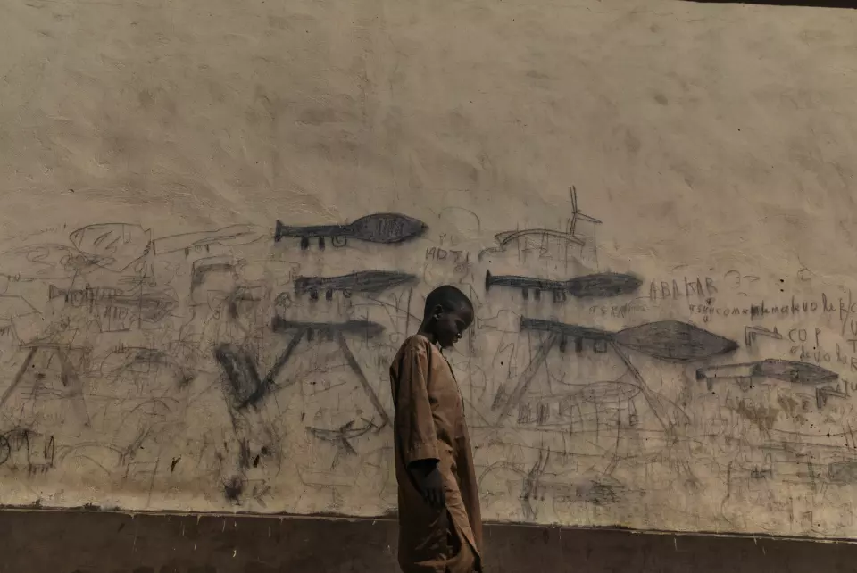 En foreldreløs gutt går forbi en vegg med tegninger av rakettdrevne granatkastere, i Bol, Tsjad. Mange foreldreløse barn, inkludert nigerianske flyktninger, bor på koranskoler og blir sendt ut for å tigge deler av dagen. Foto: Marco Gualazzini / Contrasto